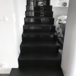 Archistone Wojciech Dodzian - Nowoczesne, czarne schody wewnętrzne z połyskiem, prowadzące do góry. Minimalistyczny design, białe ściany, drewniana podłoga. Widoczny fragment dekoracji na ścianie.