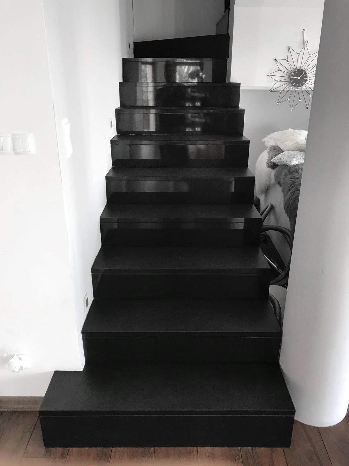 Nowoczesne, czarne schody wewnętrzne z połyskiem, prowadzące do góry. Minimalistyczny design, białe ściany, drewniana podłoga. Widoczny fragment dekoracji na ścianie.