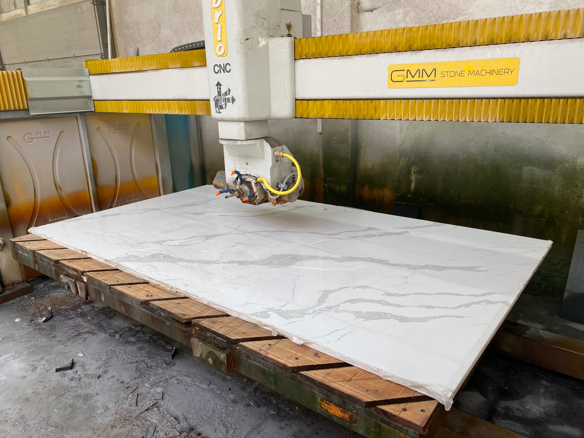 Obróbka blatu z konglomeratu na maszynie CNC. Widoczna głowica tnąca i fragment blatu z szarymi żyłkami. W tle elementy maszyny i posadzka.