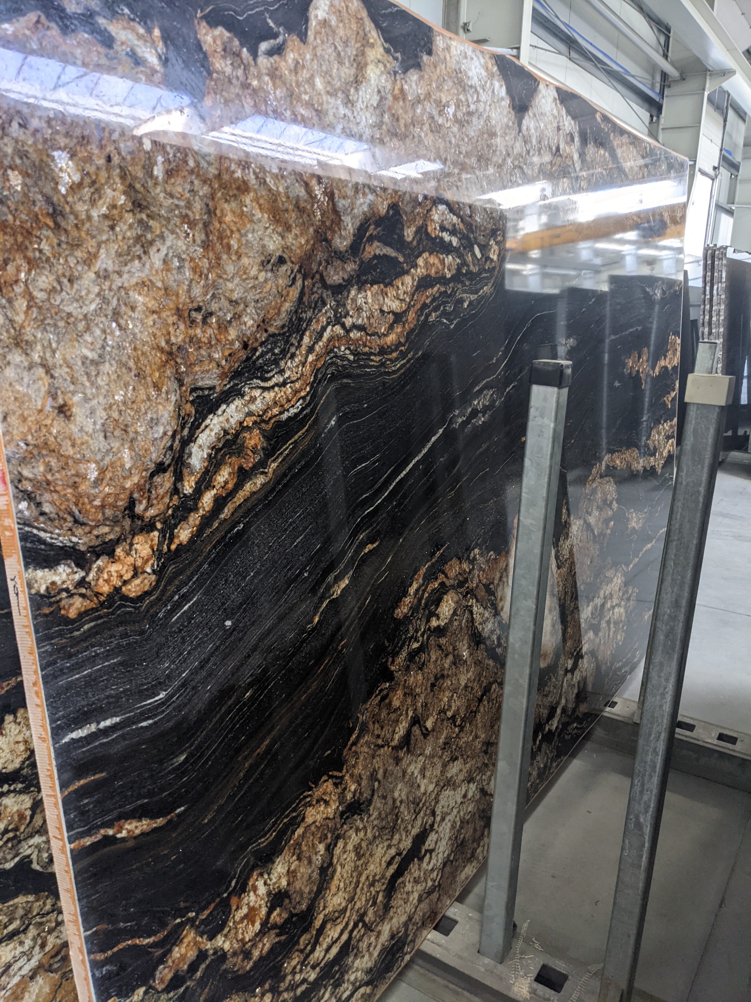 Polerowany slab kamienny o unikalnym wzorze z przewagą czerni, brązu i bieli, oparty o metalowy stojak w hali produkcyjnej. Widoczne refleksy światła.