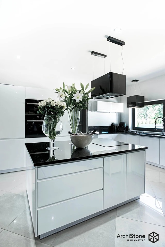 Błyszczący, czarny blat kamienny na wyspie kuchennej z białymi frontami. Na blacie wazony z białymi kwiatami. Nowoczesna kuchnia z czarnymi okapami.