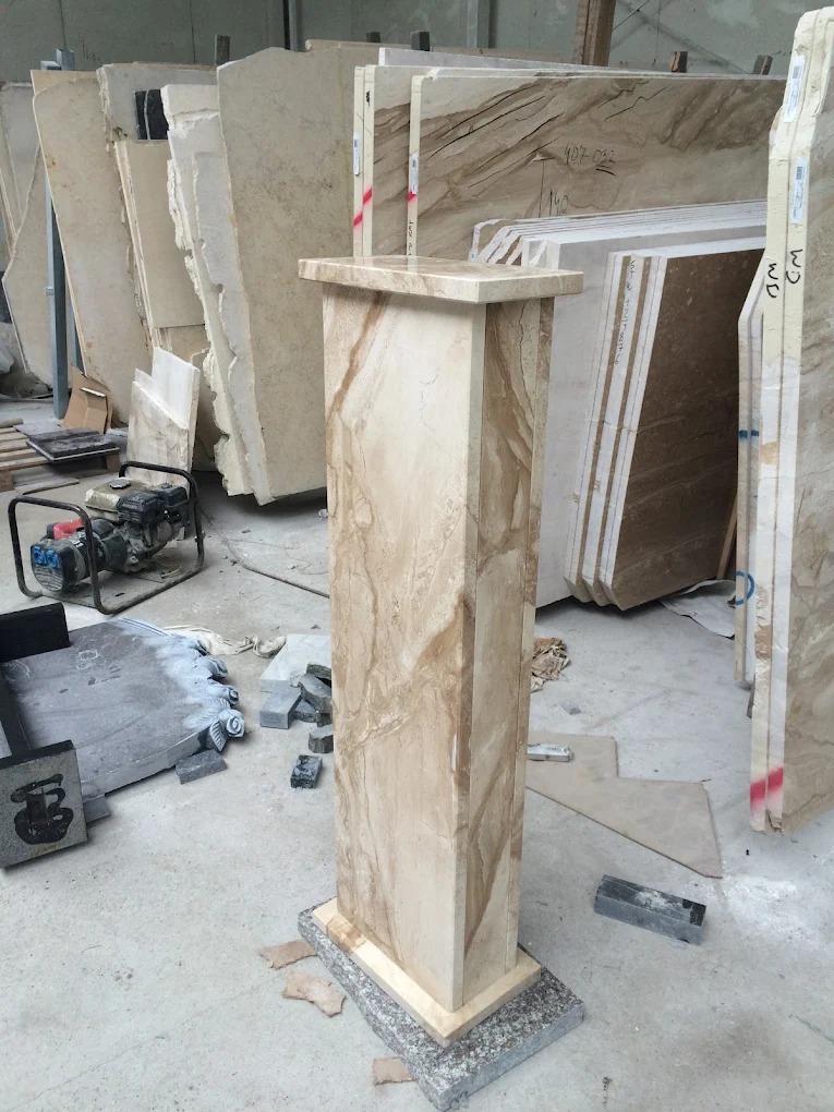 Kolumna z beżowego kamienia z wyraźnym wzorem, na tle płyt kamiennych w warsztacie. Widoczne narzędzia i elementy obróbki kamienia. Nietypowy element dekoracyjny.