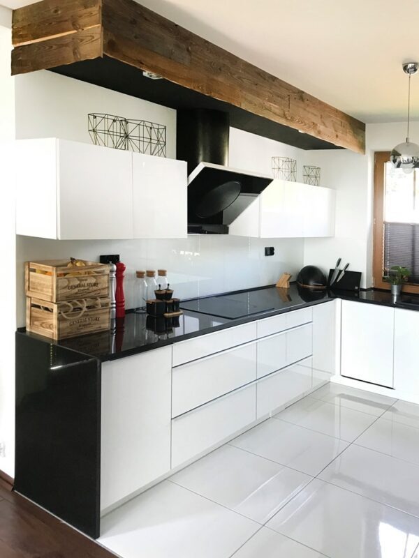 Nowoczesna kuchnia z czarnym blatem i białymi szafkami, drewniana belka stropowa, minimalistyczny design, białe płytki podłogowe, oświetlenie punktowe.
