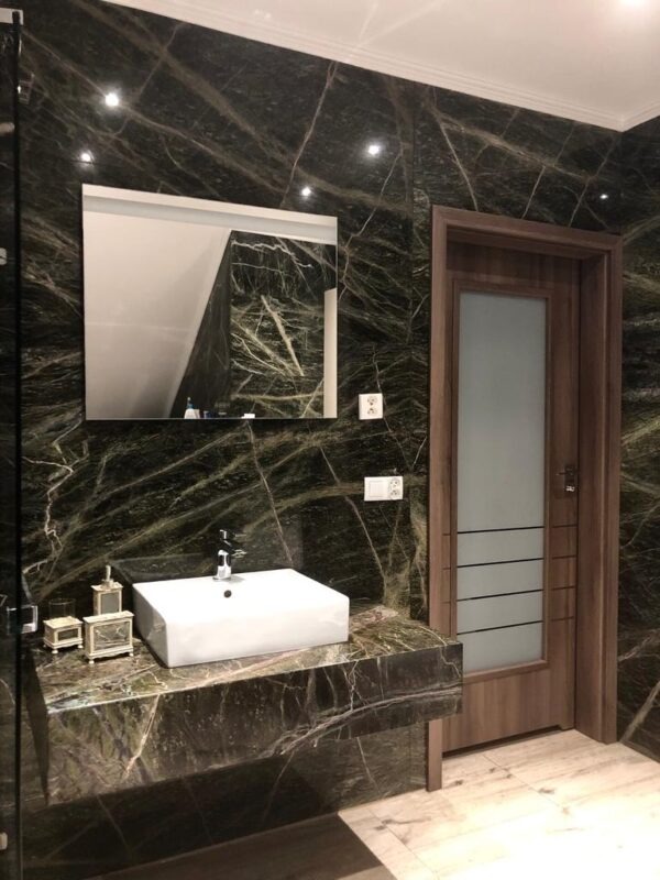 Elegancka łazienka z blatem i ścianami z ciemnego kamienia z wyraźnym żyłkowaniem. Biała umywalka nablatowa i drewniane drzwi z matowym szkłem.