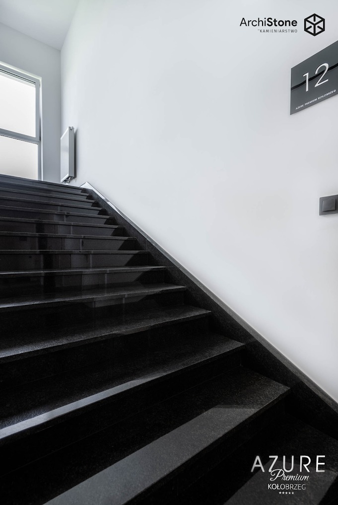 Eleganckie, czarne schody z kamienia naturalnego w nowoczesnym wnętrzu. Widoczne logo firmy kamieniarskiej w górnym rogu. Minimalistyczny design, dobre oświetlenie.