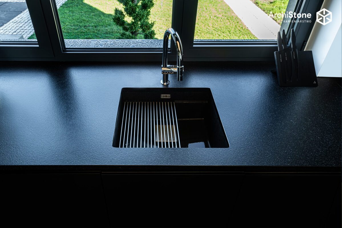 Czarny, matowy blat kamienny z wbudowanym zlewem i ociekaczem, bateria chromowana, widok na zieleń za oknem. Minimalistyczny design.