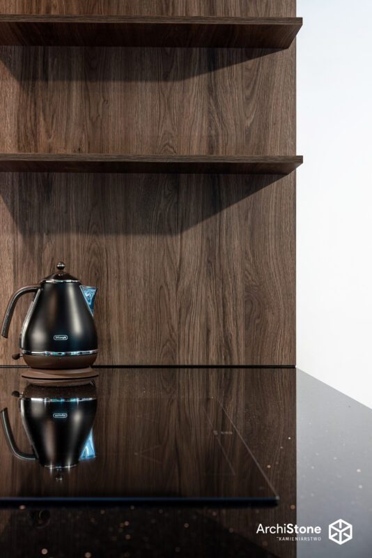 Czarny czajnik elektryczny na blacie z ciemnego kamienia z refleksami, na tle drewnianej zabudowy kuchennej z półkami. Minimalistyczny design.
