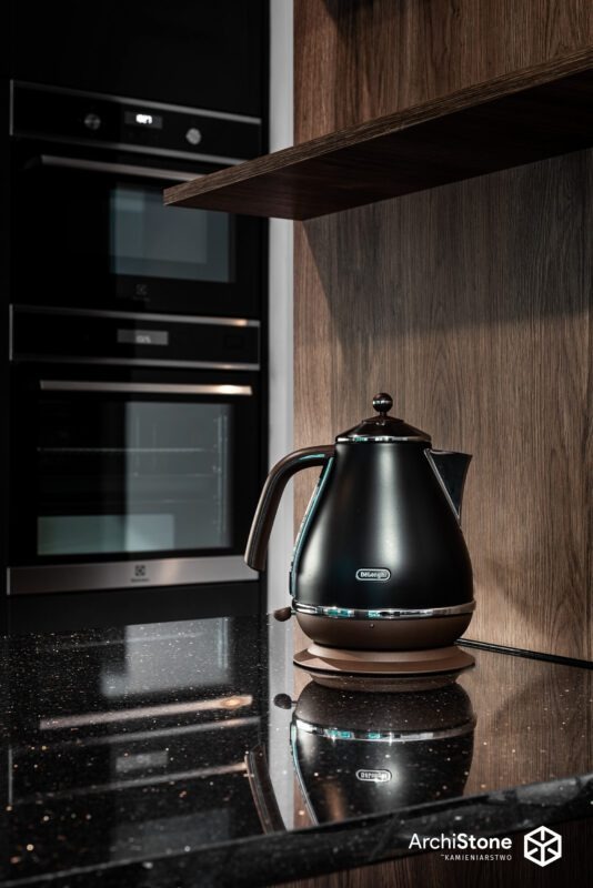 Błyszczący, czarny blat z kamienia z drobinkami, na nim czajnik Delonghi. W tle zabudowa kuchenna z piekarnikiem i drewnianą półką. Ujęcie z niskiego kąta.