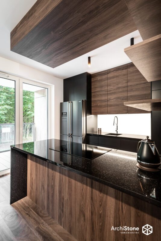 Czarny, błyszczący blat z drobnymi, jasnymi inkluzjami, na tle nowoczesnej kuchni z drewnianymi i czarnymi szafkami. Widoczny fragment płyty grzewczej i czajnik.