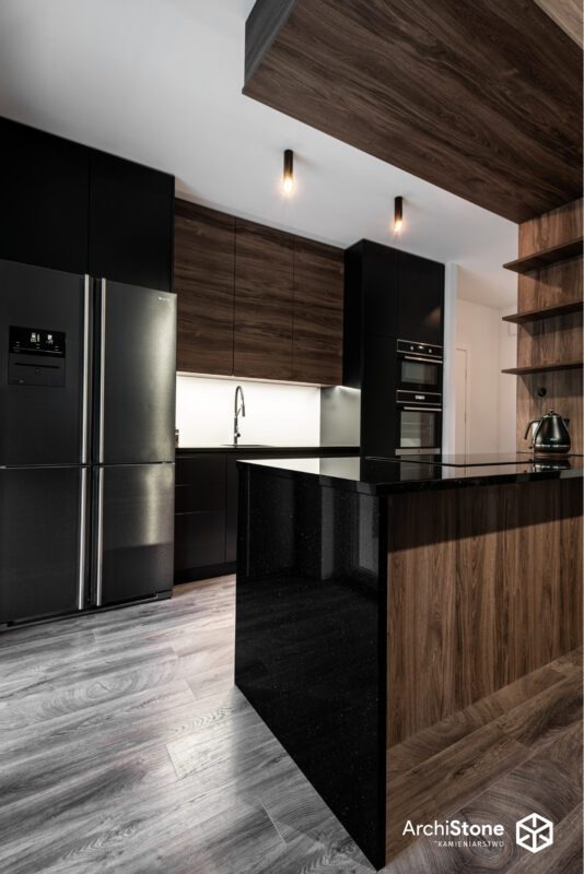 Nowoczesna kuchnia z czarnym blatem wyspy, drewnianymi szafkami i podłogą w odcieniach szarości. Minimalistyczne oświetlenie i sprzęt AGD w zabudowie.