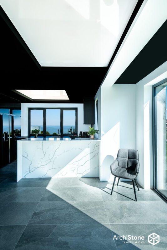 Nowoczesna kuchnia w Koszalinie z blatem z konglomeratu o wzorze marmuru, szare płytki podłogowe i czarne elementy sufitu. Minimalistyczny design i naturalne światło.