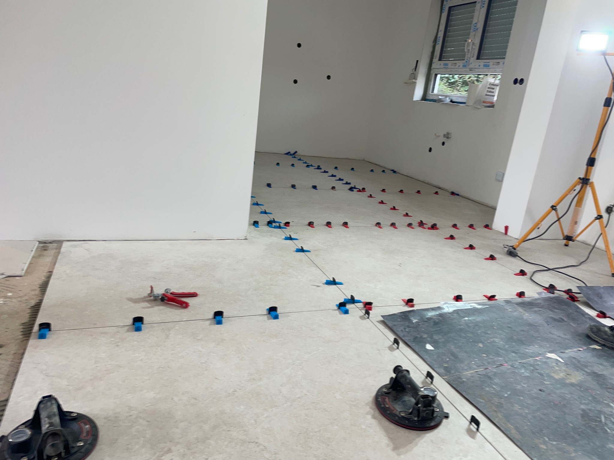 Układanie płytek podłogowych z użyciem systemu poziomowania; widoczne przyssawki, szczypce i lampa budowlana. Jasne płytki, niebiesko-czerwone kliny.