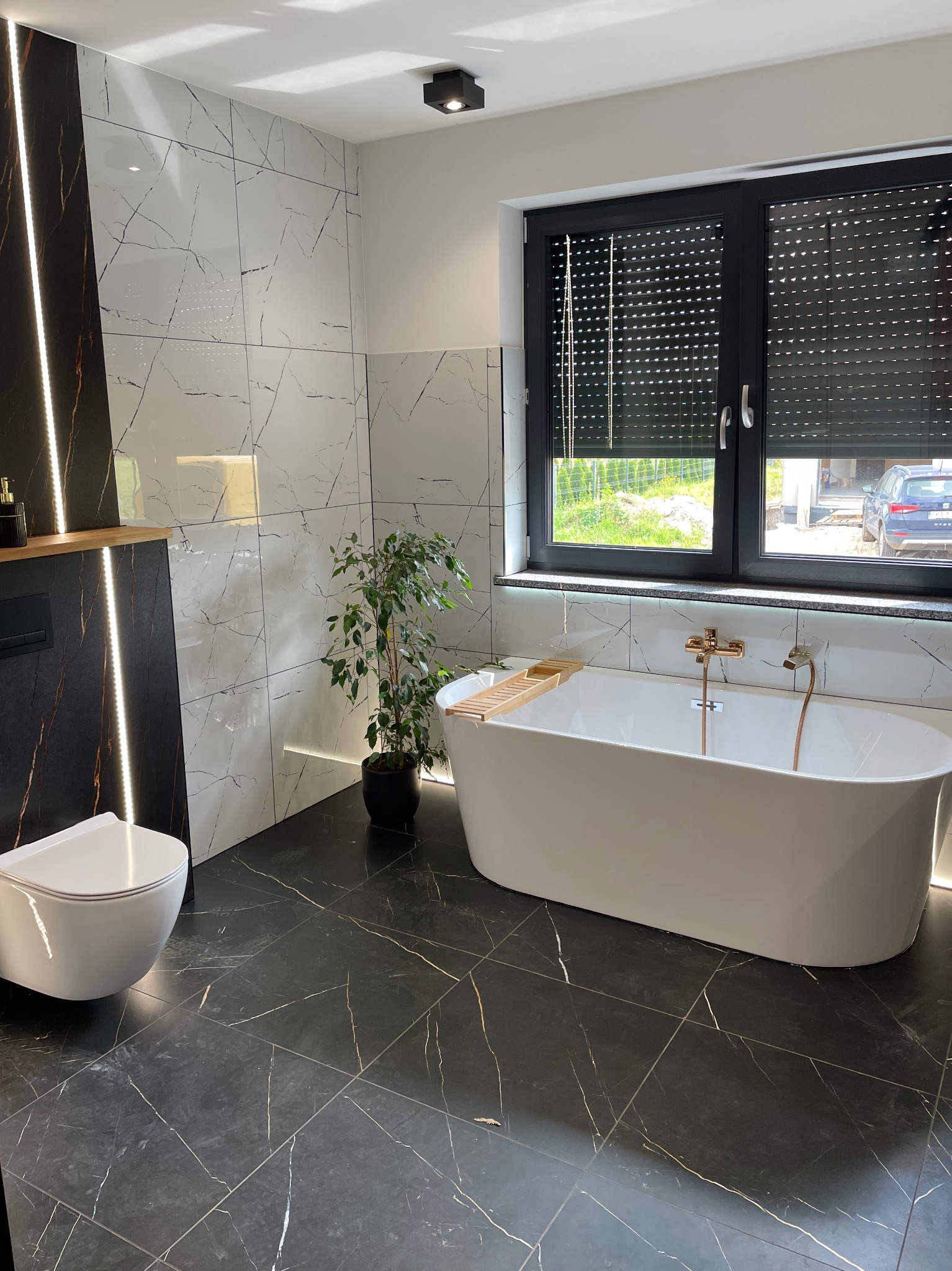 Nowoczesna łazienka z czarno-białymi płytkami imitującymi marmur, wolnostojącą wanną i podświetleniem LED. Minimalistyczny design z rośliną doniczkową.