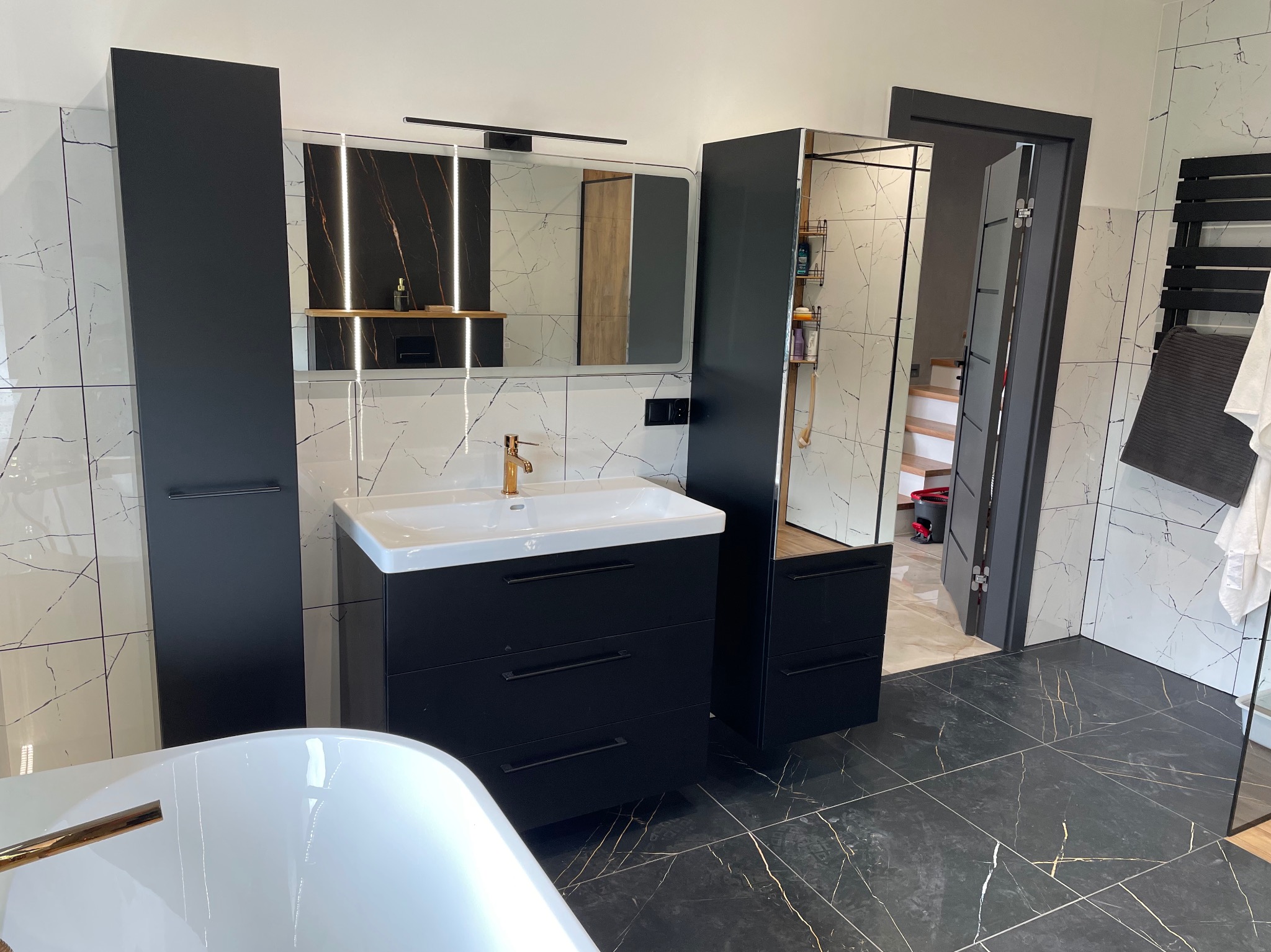 Nowoczesna łazienka z czarnymi meblami, białymi płytkami imitującymi marmur i złotą armaturą. Widoczna wanna, umywalka z szafką i wysoka szafka.