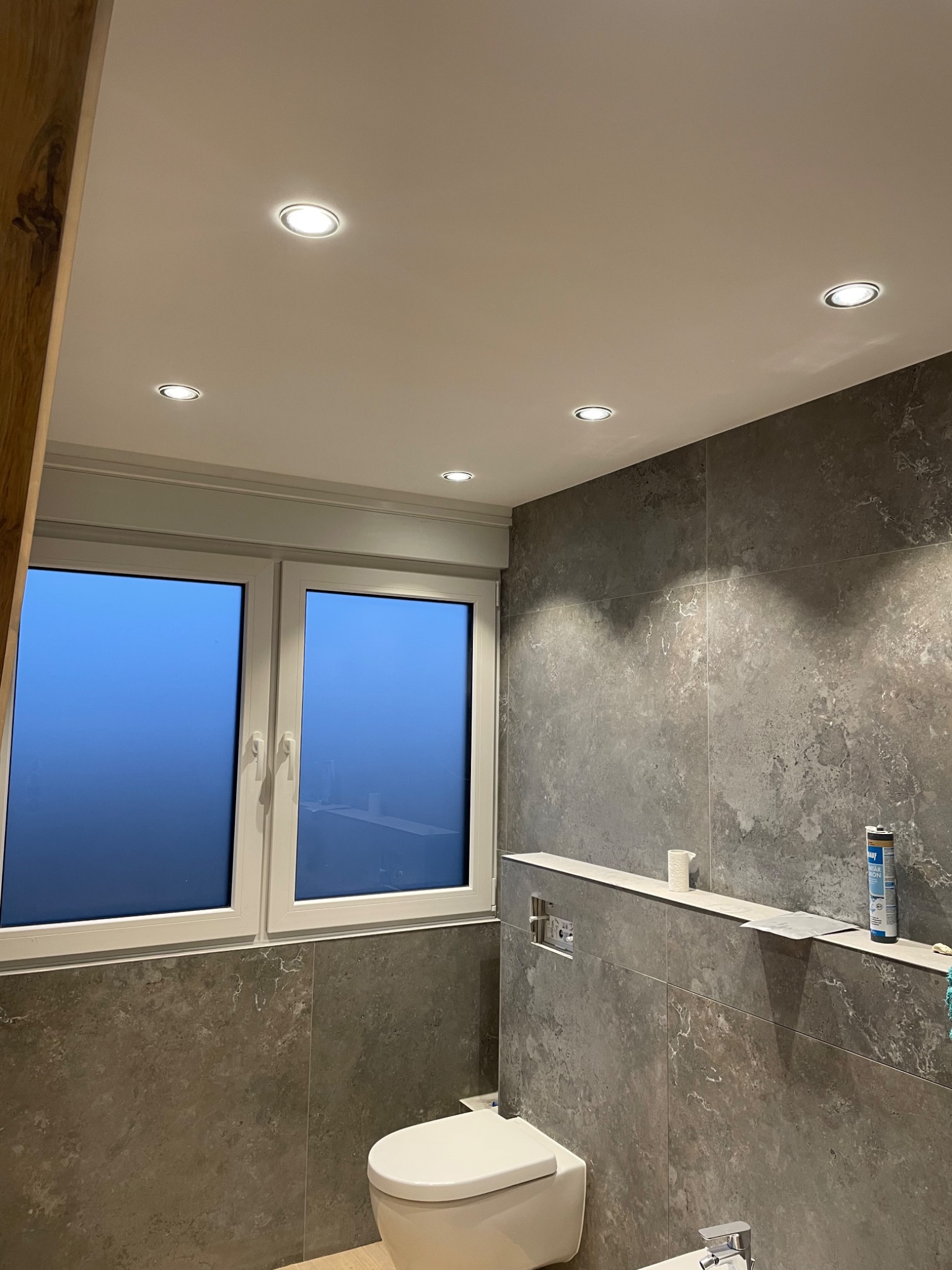 Wykończona łazienka z szarymi płytkami imitującymi kamień, podwieszaną toaletą i oknem. Sufit z oświetleniem punktowym. Widoczny fragment drewnianej ramy.