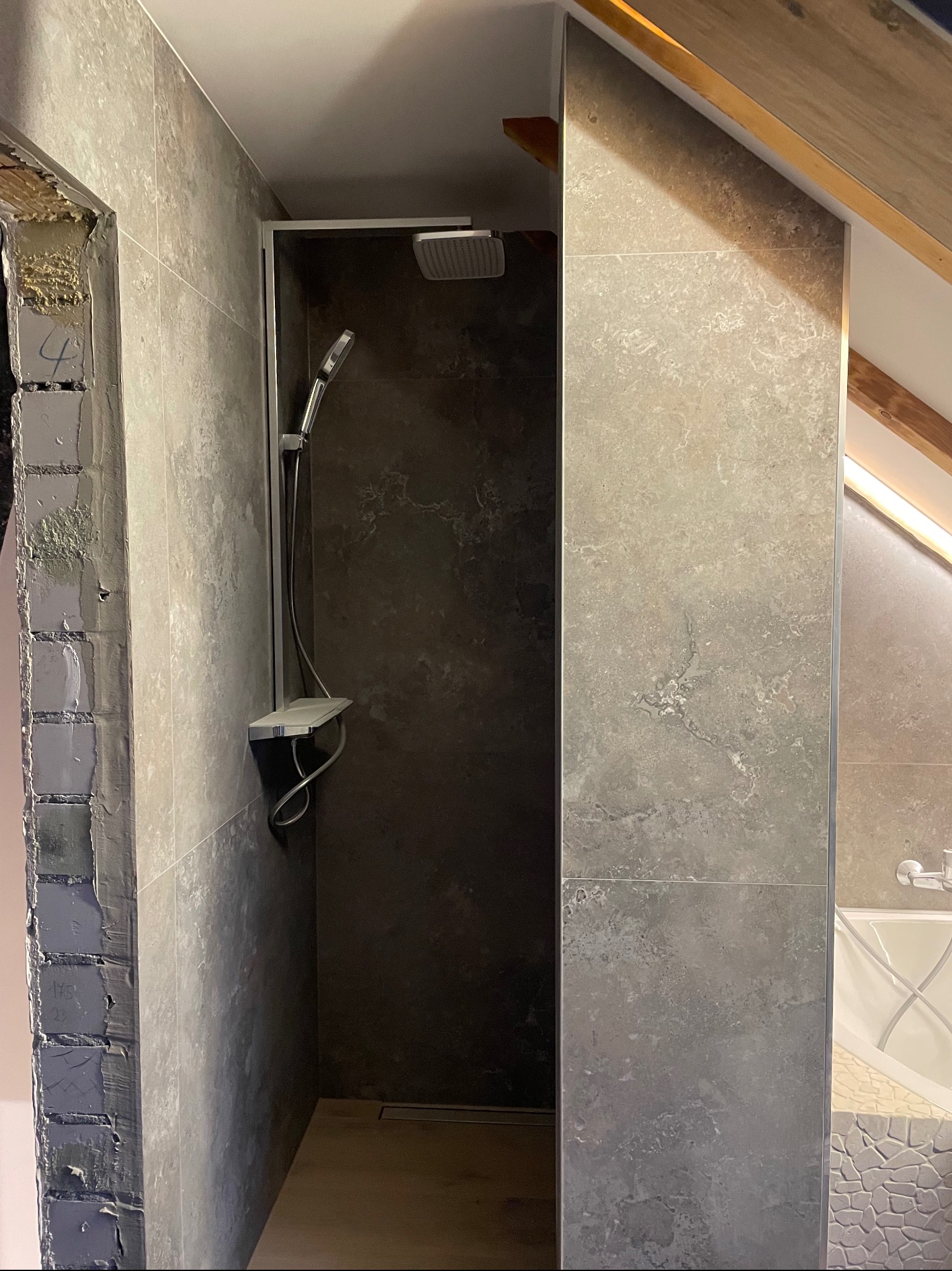 Wykończona kabina prysznicowa z szarymi płytkami w łazience na poddaszu, widoczny fragment wanny i surowy mur po lewej stronie.