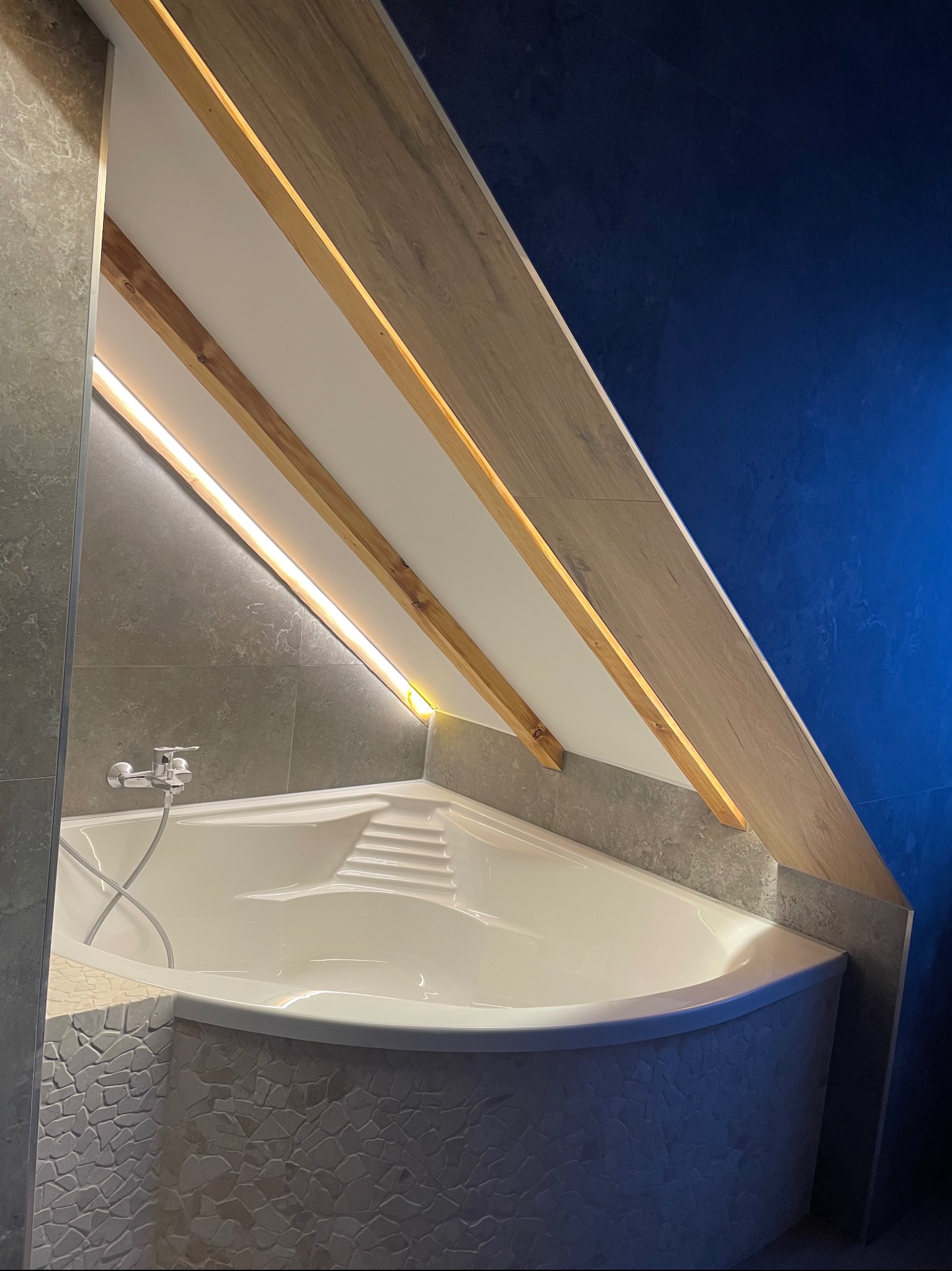 Nowoczesna łazienka na poddaszu z wanną obudowaną mozaiką, oświetleniem LED i szarą glazurą. Niebieska ściana dodaje kontrastu.