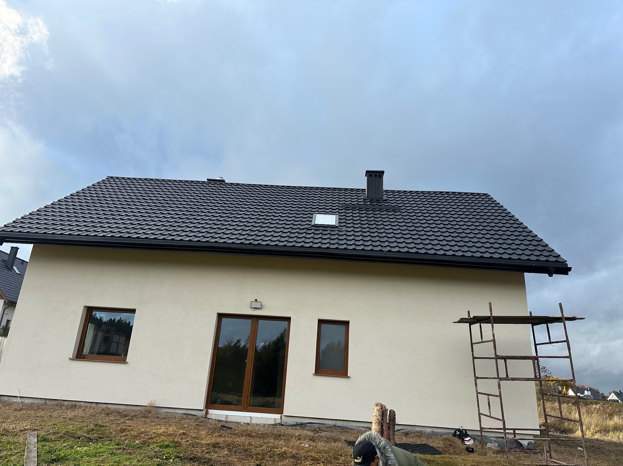 Dom jednorodzinny z ciemną dachówką, widoczny fragment rusztowania. Dach z oknem dachowym i kominem. Pochmurne niebo w tle.