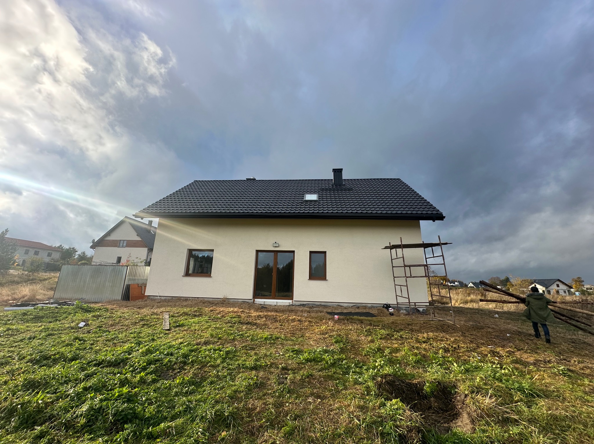Dom jednorodzinny z ciemną dachówką na tle pochmurnego nieba. Widoczne rusztowanie i osoba przenosząca drewno, co sugeruje prace wykończeniowe.