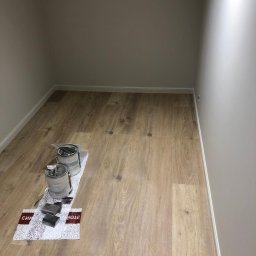 METR2 - Pomalowane na szaro wnętrze z drewnianą podłogą. Na podłodze pędzle i dwie puszki z farbą na folii ochronnej. Świeżo po malowaniu.