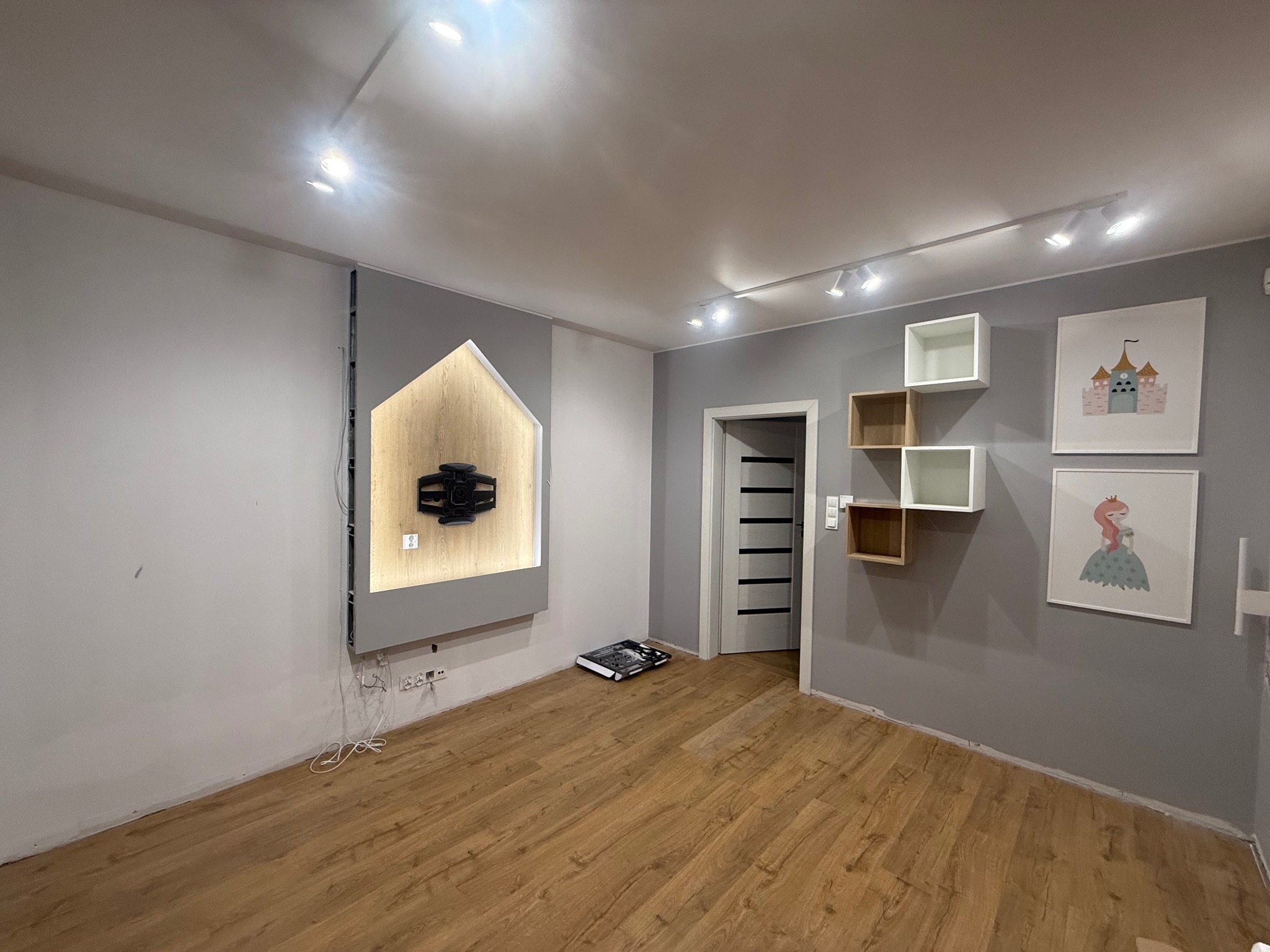 Jasne wnętrze pokoju po remoncie: drewniana podłoga, szara ściana z podświetloną wnęką na TV w kształcie domku, półki i obrazki na ścianie. Nowoczesne oświetlenie.
