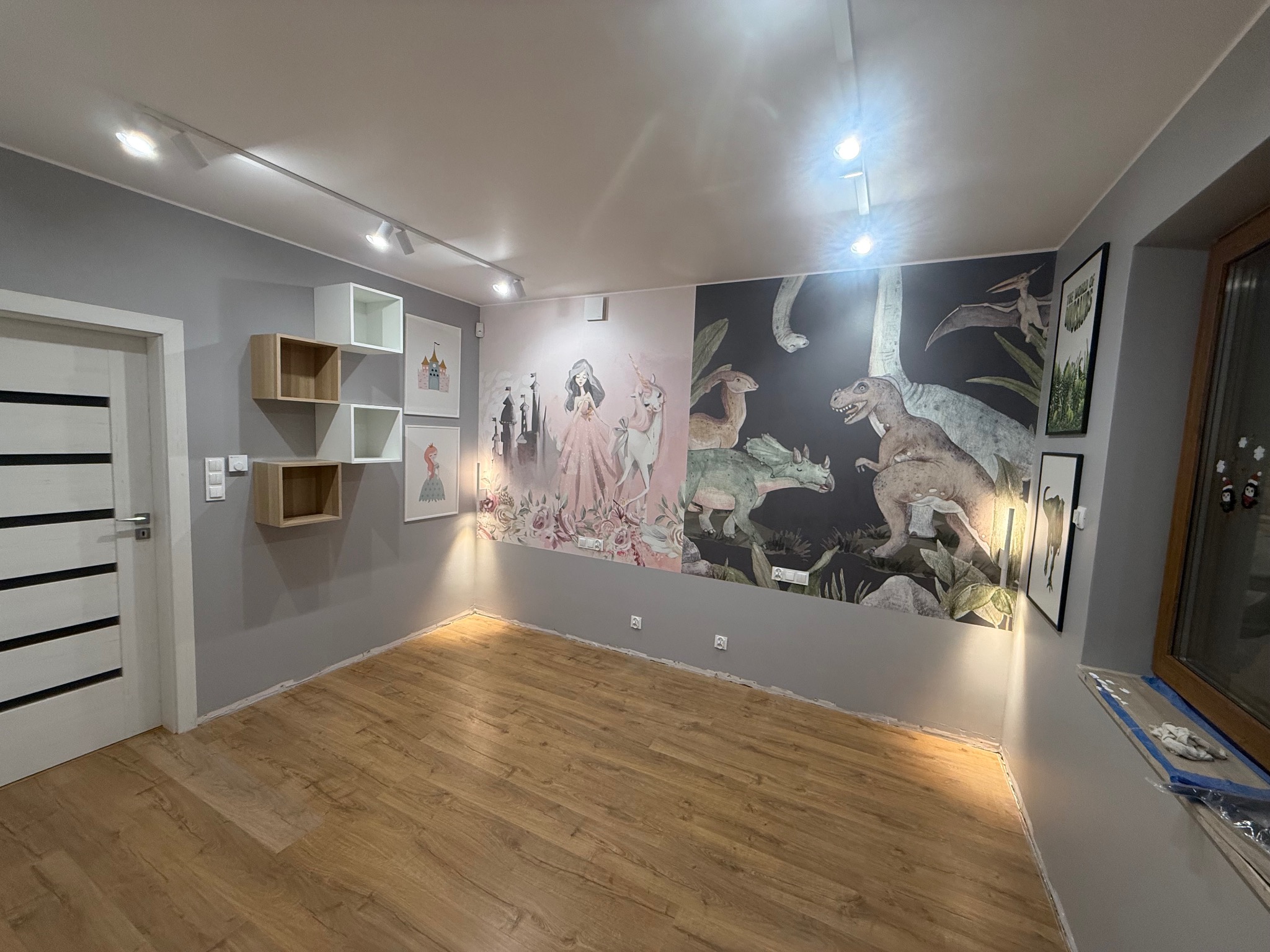 Pokój dziecięcy po malowaniu ścian z motywem bajkowym i dinozaurów. Na ścianie półki i obrazy. Podłoga drewniana. Oświetlenie sufitowe punktowe.