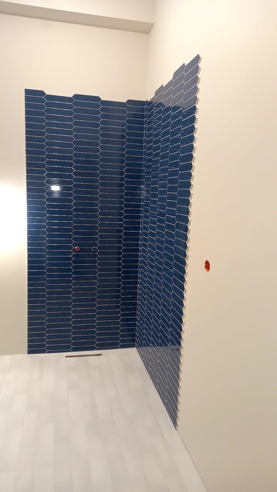 Niebieska mozaika heksagonalna na ścianie kabiny prysznicowej, białe płytki podłogowe, minimalistyczny design, widoczne przyłącza wodne.