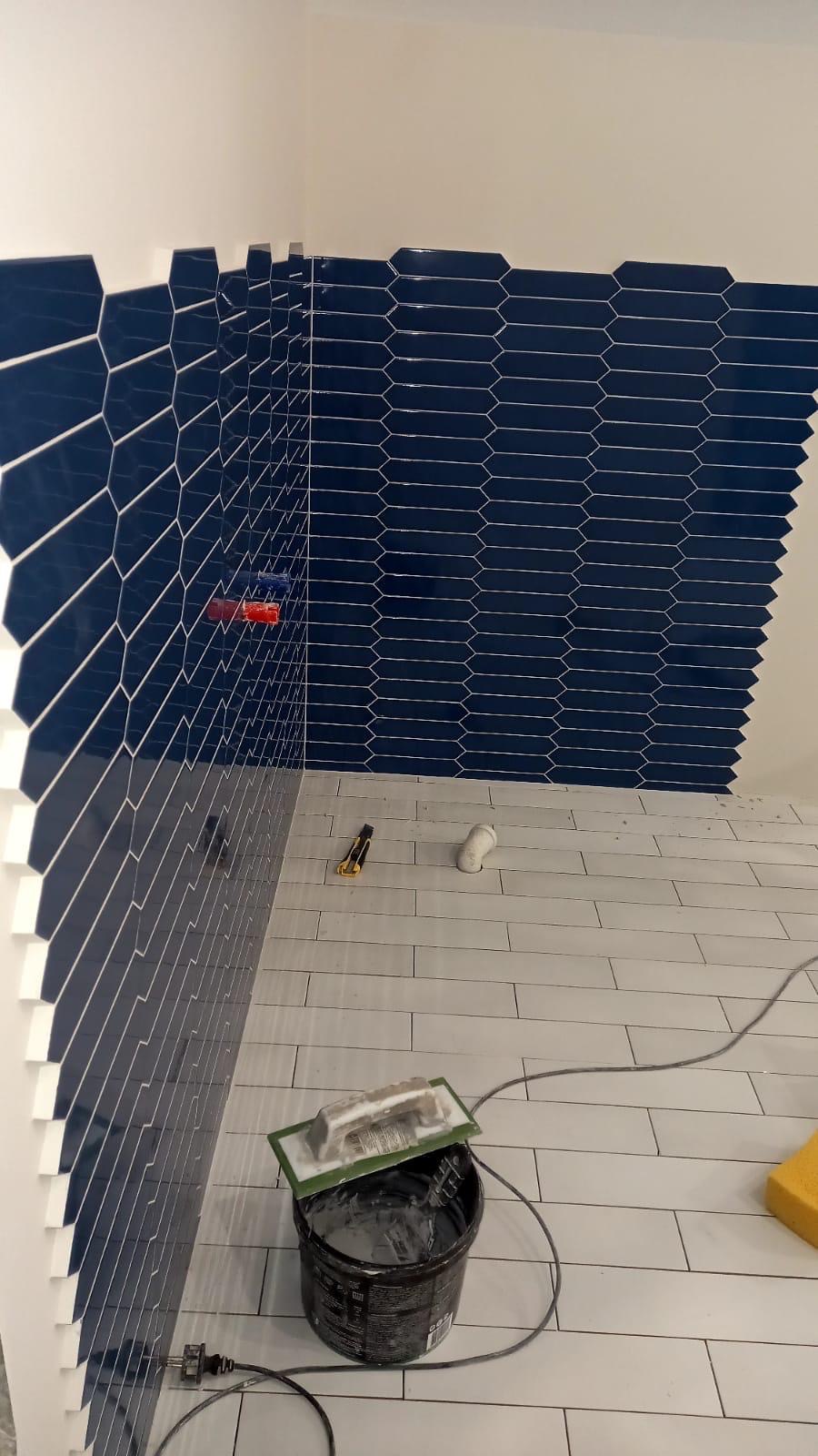 Łazienka w trakcie wykańczania: ułożone ciemnoniebieskie, podłużne płytki na ścianie i jasne płytki podłogowe. Widoczne narzędzia glazurnicze i wiadro z zaprawą.