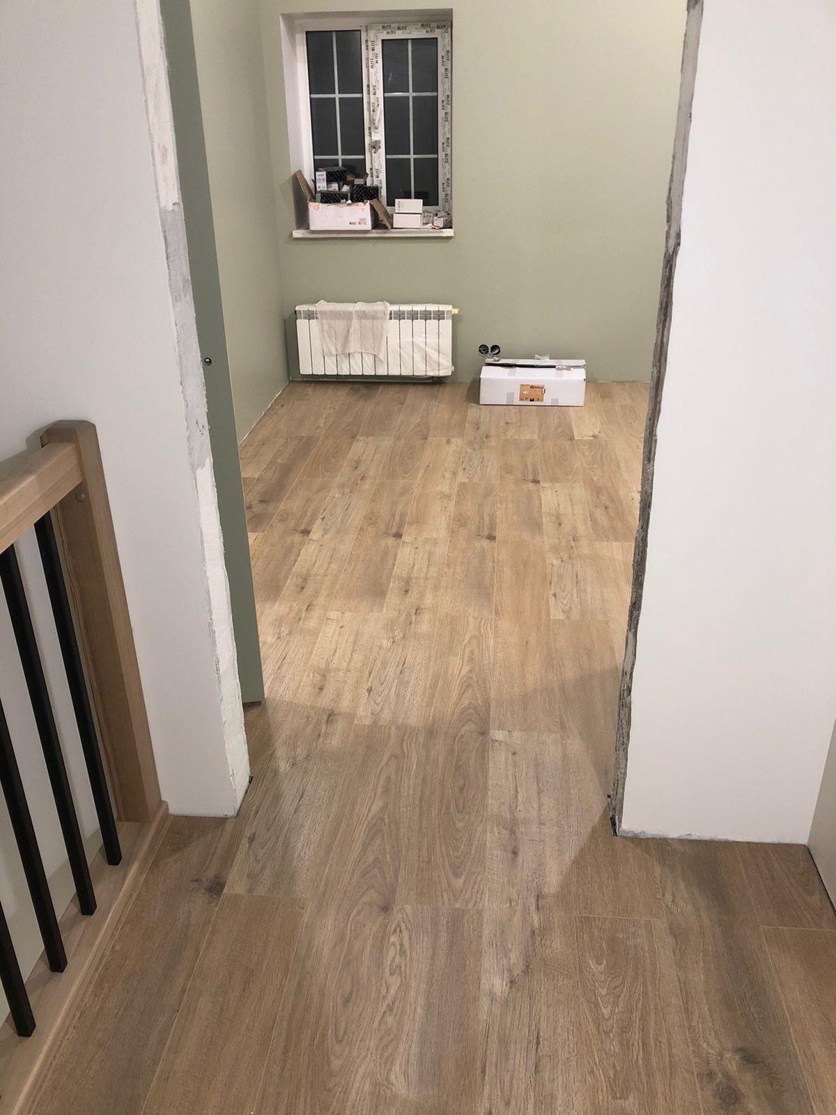 Jasne panele podłogowe ułożone w pokoju z oknem i grzejnikiem. Widoczne fragmenty ścian w odcieniach bieli i zieleni. Częściowo widoczna balustrada schodów.
