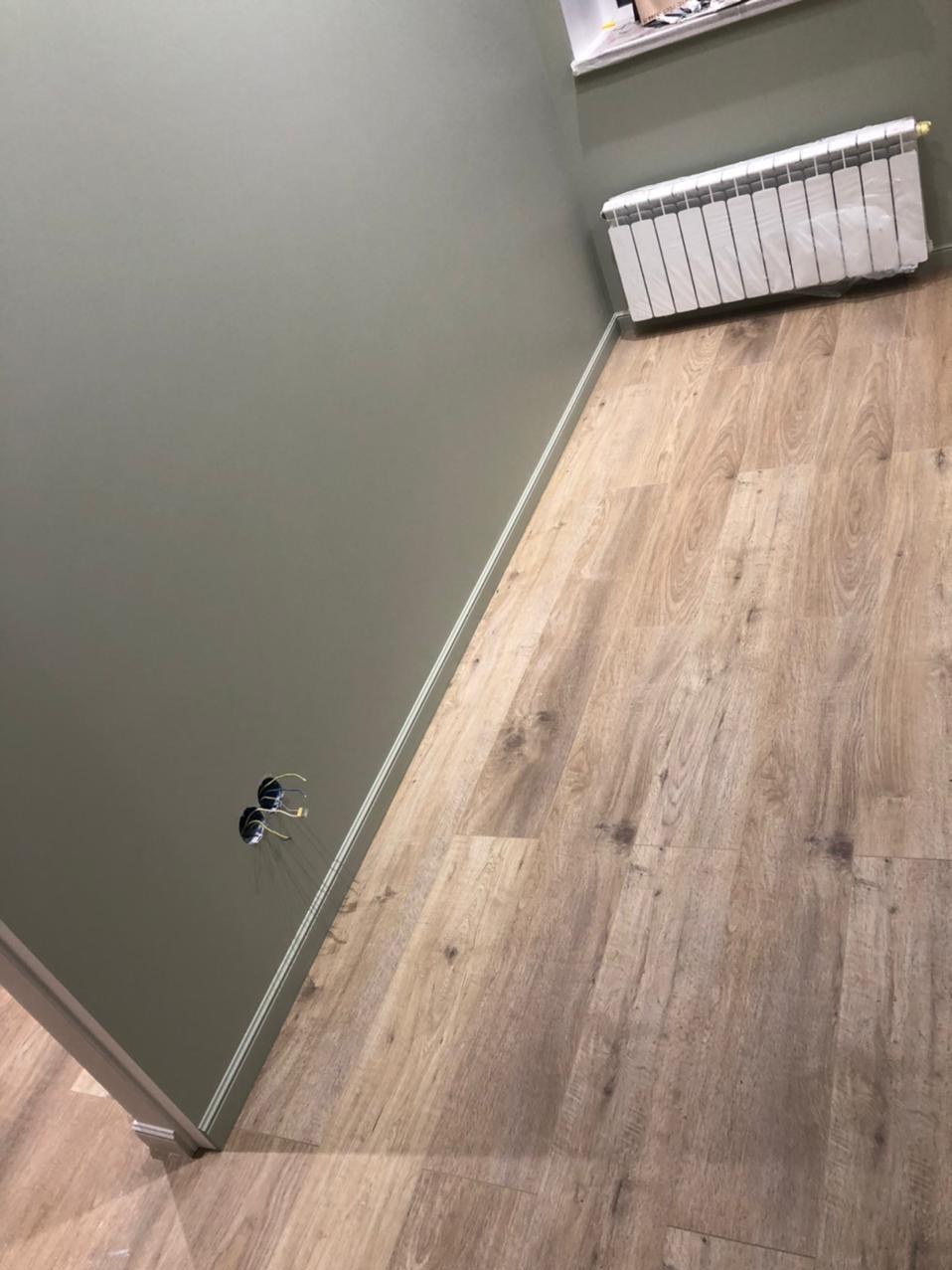 Ściana w odcieniach zieleni z gniazdkami elektrycznymi, drewnopodobna podłoga, biały grzejnik pod oknem. Wykończenie wnętrza w neutralnej kolorystyce.