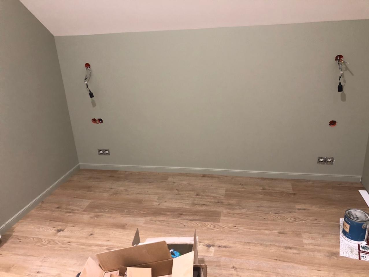 Pokój po malowaniu ścian na oliwkowy odcień, z nową podłogą z paneli. Widoczne puszki elektryczne i wystające kable. Karton i puszka farby w rogu.