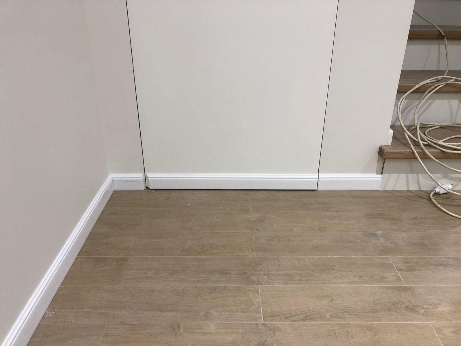 Wykończona podłoga z paneli imitujących drewno, białe listwy przypodłogowe przy ścianie i zabudowie, fragment schodów z przewodem elektrycznym w tle.
