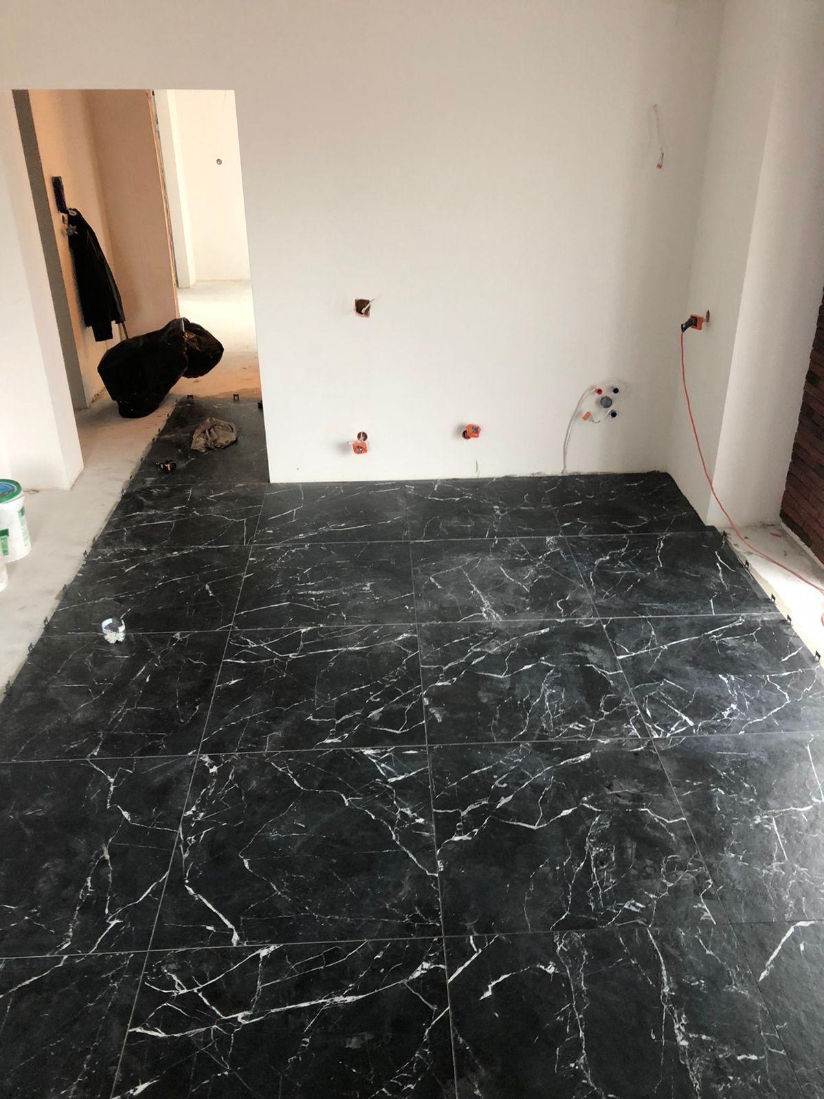 Czarne płytki imitujące marmur, ułożone na podłodze w trakcie prac wykończeniowych. Widoczne elementy instalacji elektrycznej i sanitarnej na ścianie.