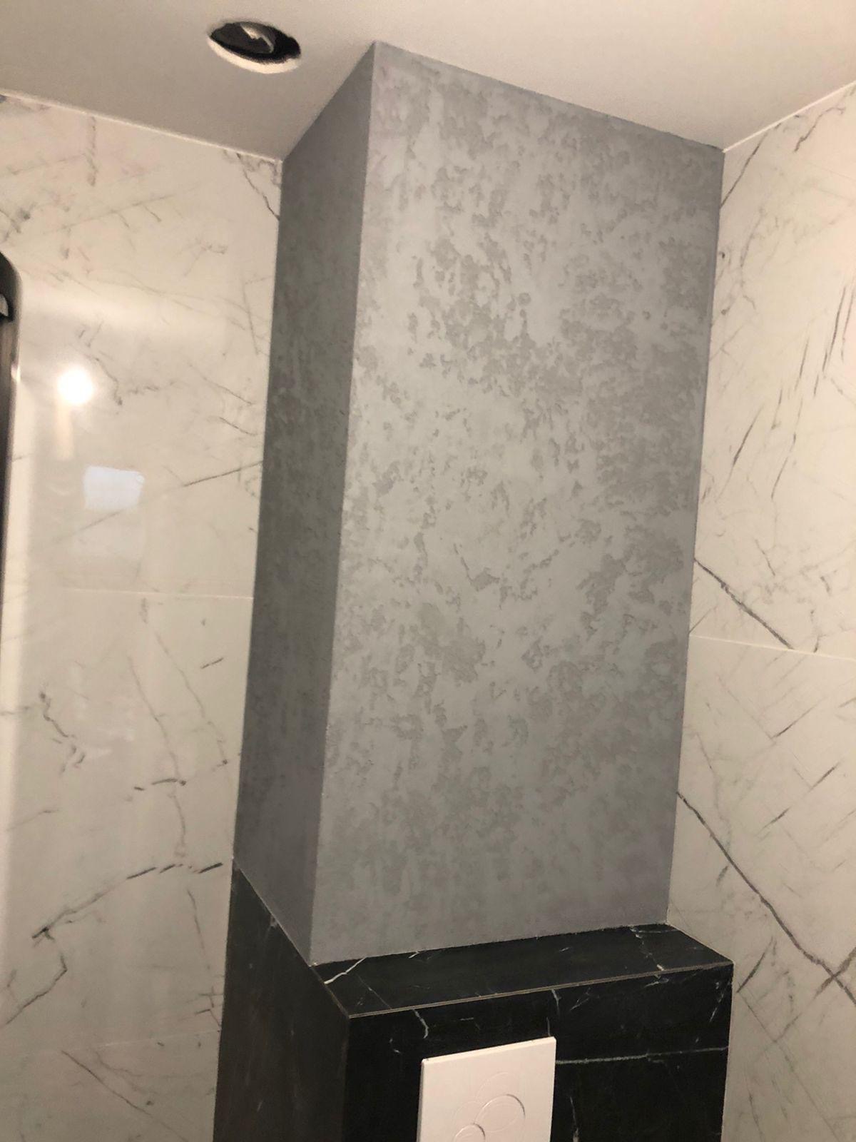 Dekoracyjny filar w łazience z efektem betonu architektonicznego, obok marmurowe płytki. Czarny cokół z przyciskiem spłuczki. Nowoczesne wykończenie.