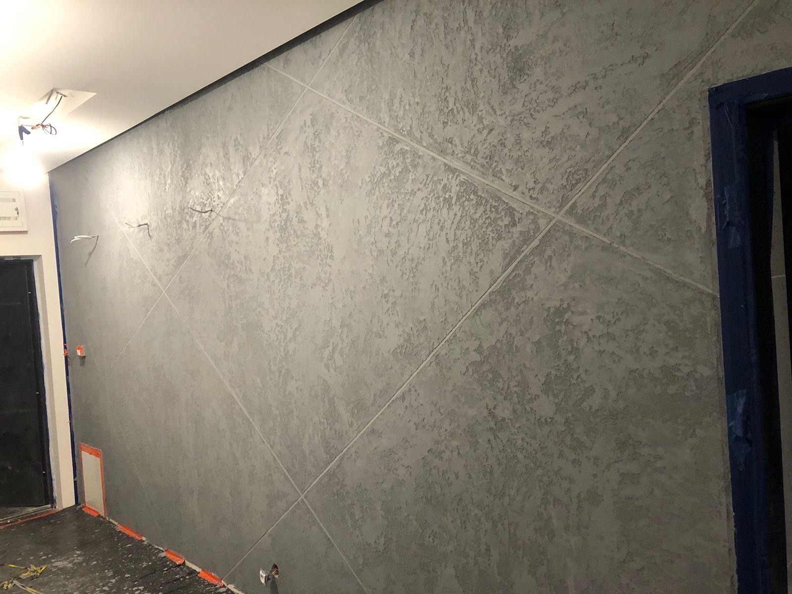 Szara ściana z dekoracyjnym efektem betonu i geometrycznym wzorem. Widoczne zabezpieczenia na podłodze i futrynie, sugerujące świeżo zakończone prace wykończeniowe.