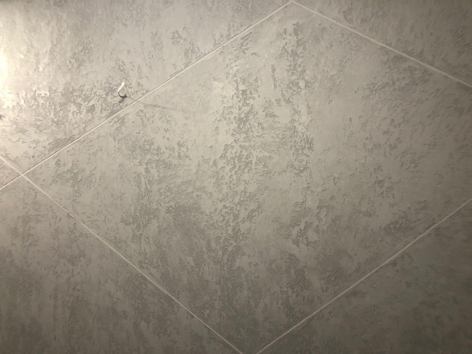 Szara ściana wykończona dekoracyjnym tynkiem strukturalnym, ułożona z płytek w romby. Widoczny haczyk na ścianie.