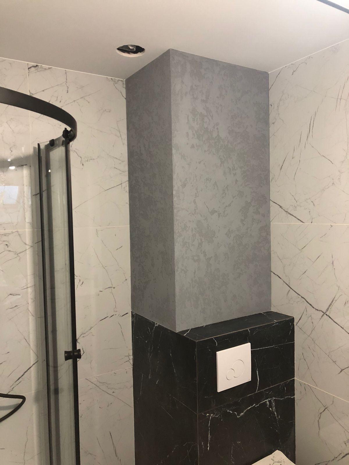 Wykończona kolumna w łazience z efektem dekoracyjnym na szarej powierzchni i czarnym marmurze u dołu, obok kabiny prysznicowej i płytek imitujących marmur.