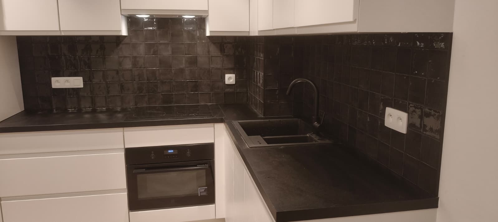 Nowoczesna kuchnia z czarnymi płytkami i blatem, białe szafki, czarna armatura. Minimalistyczny design, oświetlenie punktowe, gładkie powierzchnie.