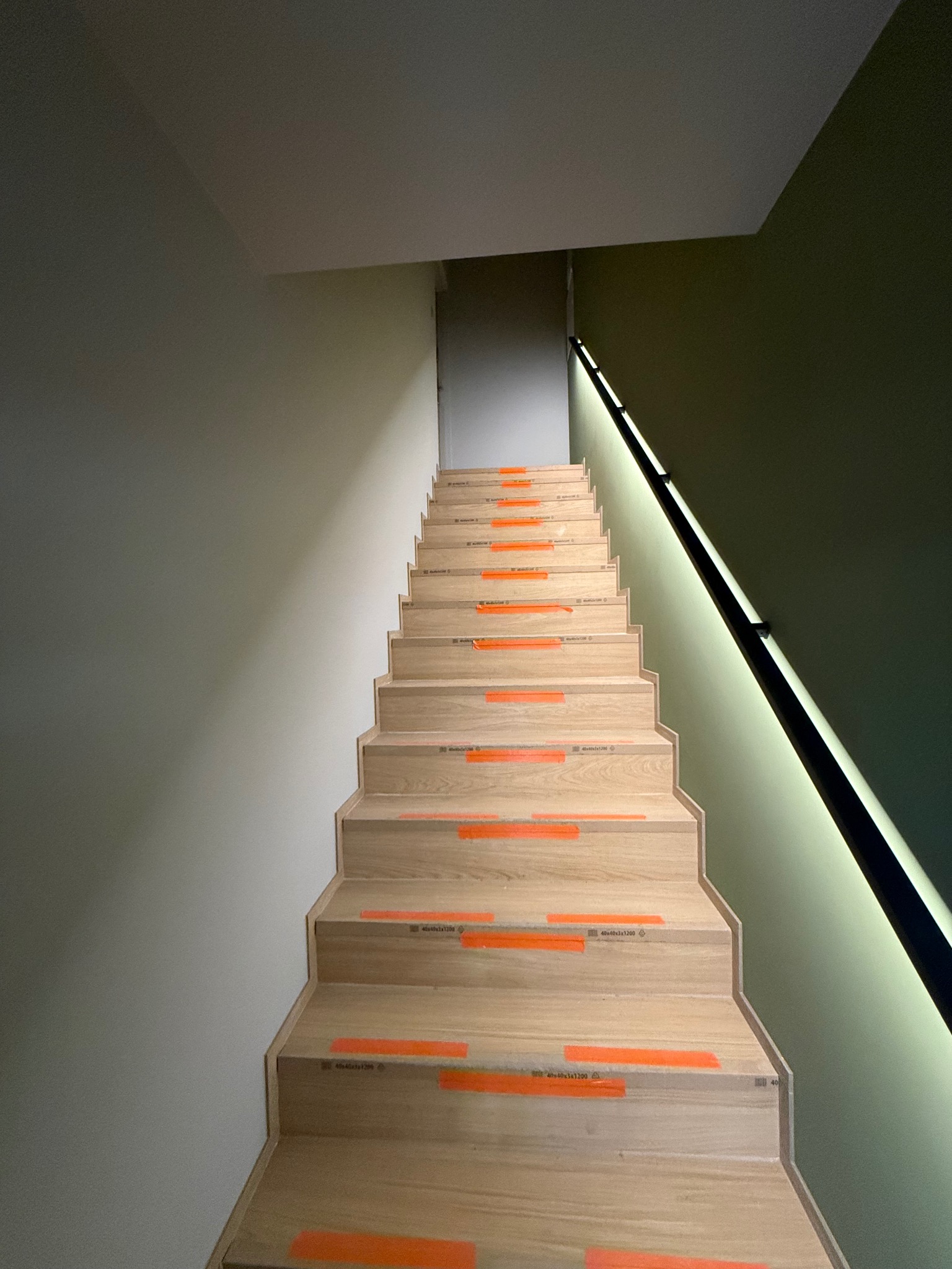 Schody drewniane z pomarańczowymi zabezpieczeniami w Łodzi, prowadzące w górę, oświetlone listwą LED przy ścianie. Widok z dołu, nowoczesne wykończenie.