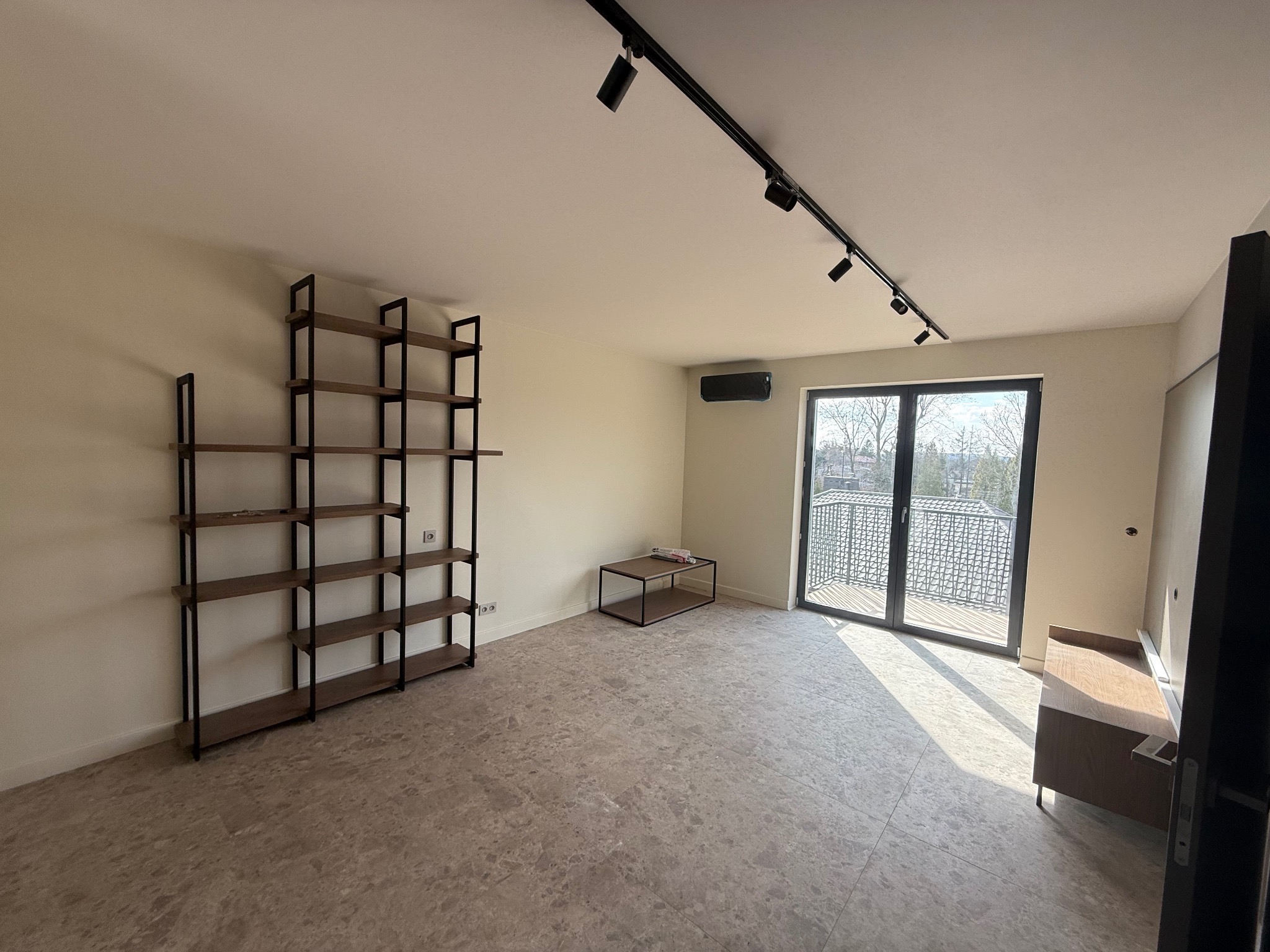 Jasne wnętrze z regałem, stolikiem i wyjściem na balkon. Podłoga wyłożona płytkami imitującymi kamień. Oświetlenie szynowe na suficie. Minimalistyczny design.