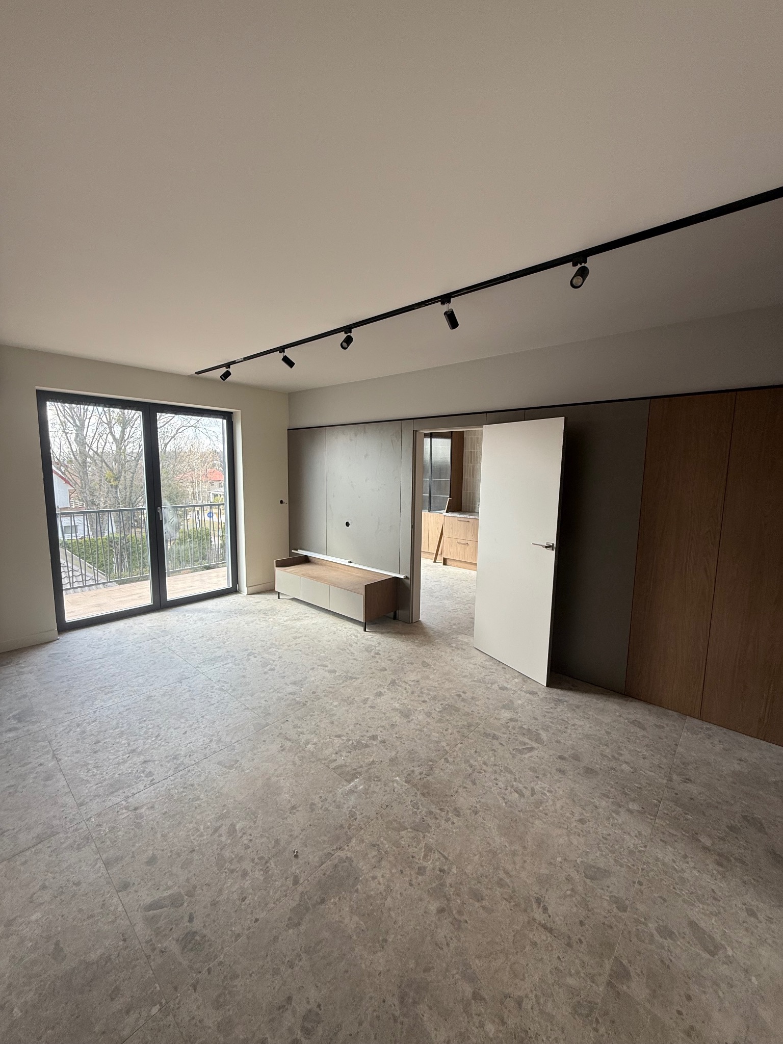 Wykończone wnętrze pokoju z szarą podłogą z płytek, oświetleniem szynowym i zabudową meblową w odcieniach szarości i drewna. Widok na balkon i otwarte drzwi do kuchni.