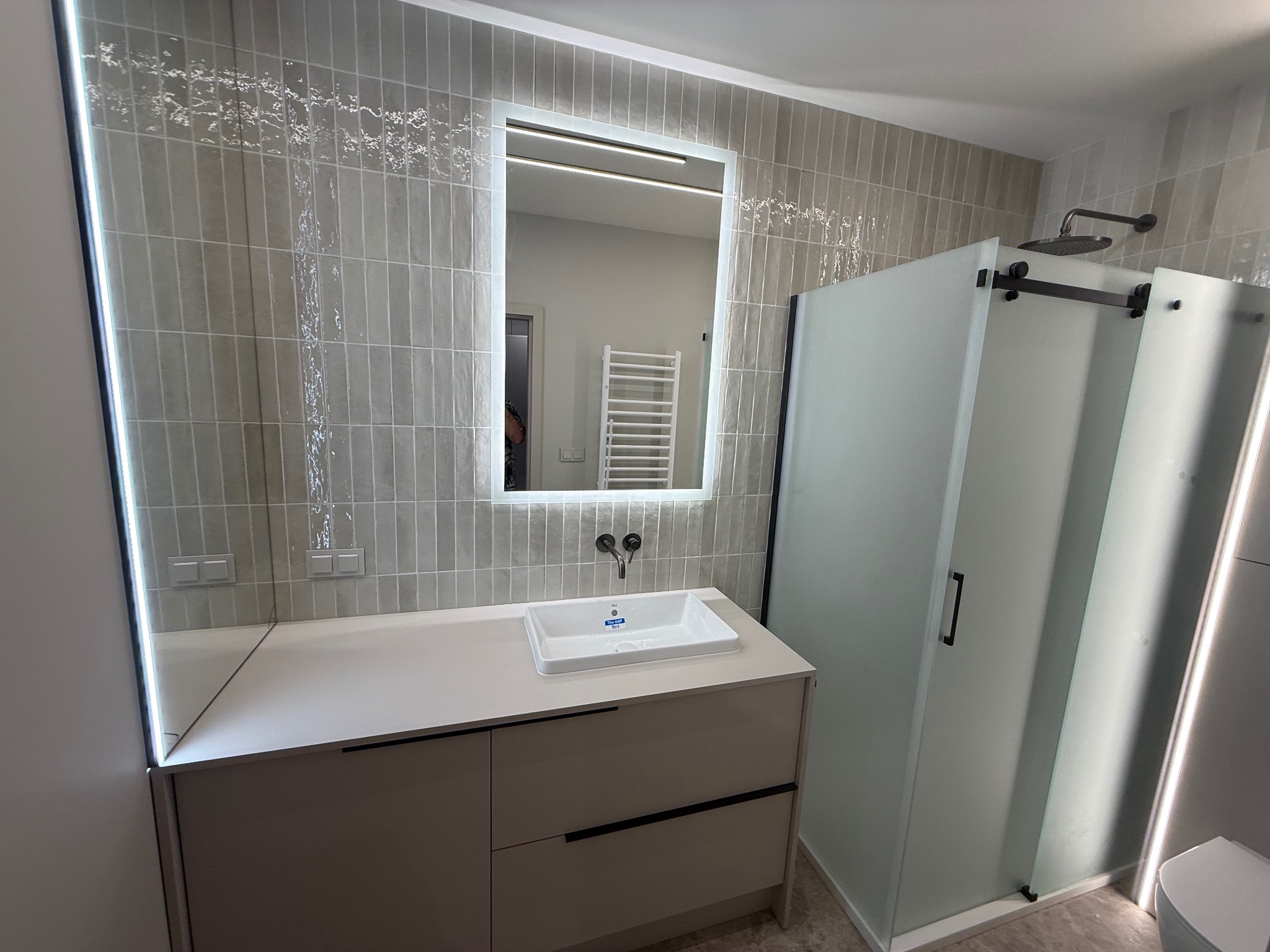 Nowoczesna łazienka z beżowymi płytkami, podświetlanym lustrem i kabiną prysznicową z przesuwnymi drzwiami. Minimalistyczna szafka pod umywalką z ciemnymi uchwytami.