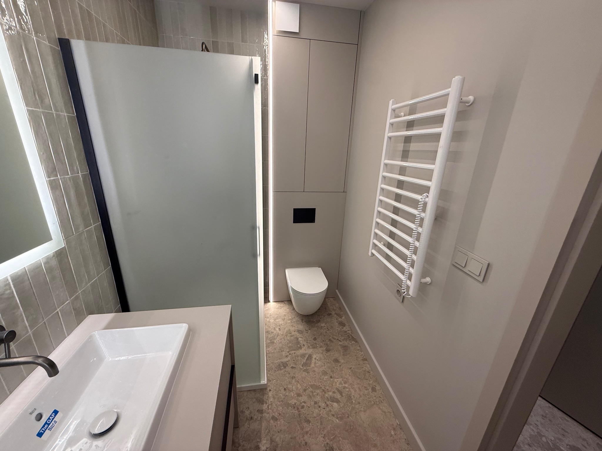 Nowoczesna łazienka w odcieniach beżu z kabiną prysznicową, umywalką, toaletą i grzejnikiem drabinkowym. Minimalistyczny design, płytki imitujące kamień na podłodze.