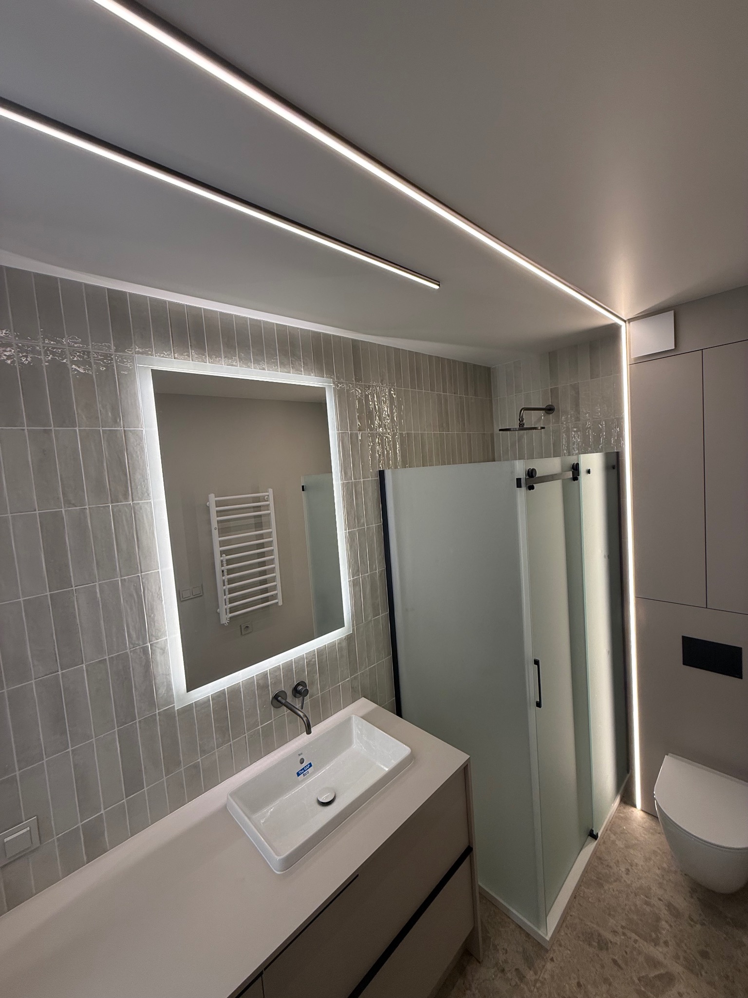 Nowoczesna łazienka w odcieniach szarości z podświetlanym lustrem, kabiną prysznicową typu walk-in i umywalką nablatową. Minimalistyczny design z akcentami świetlnymi.