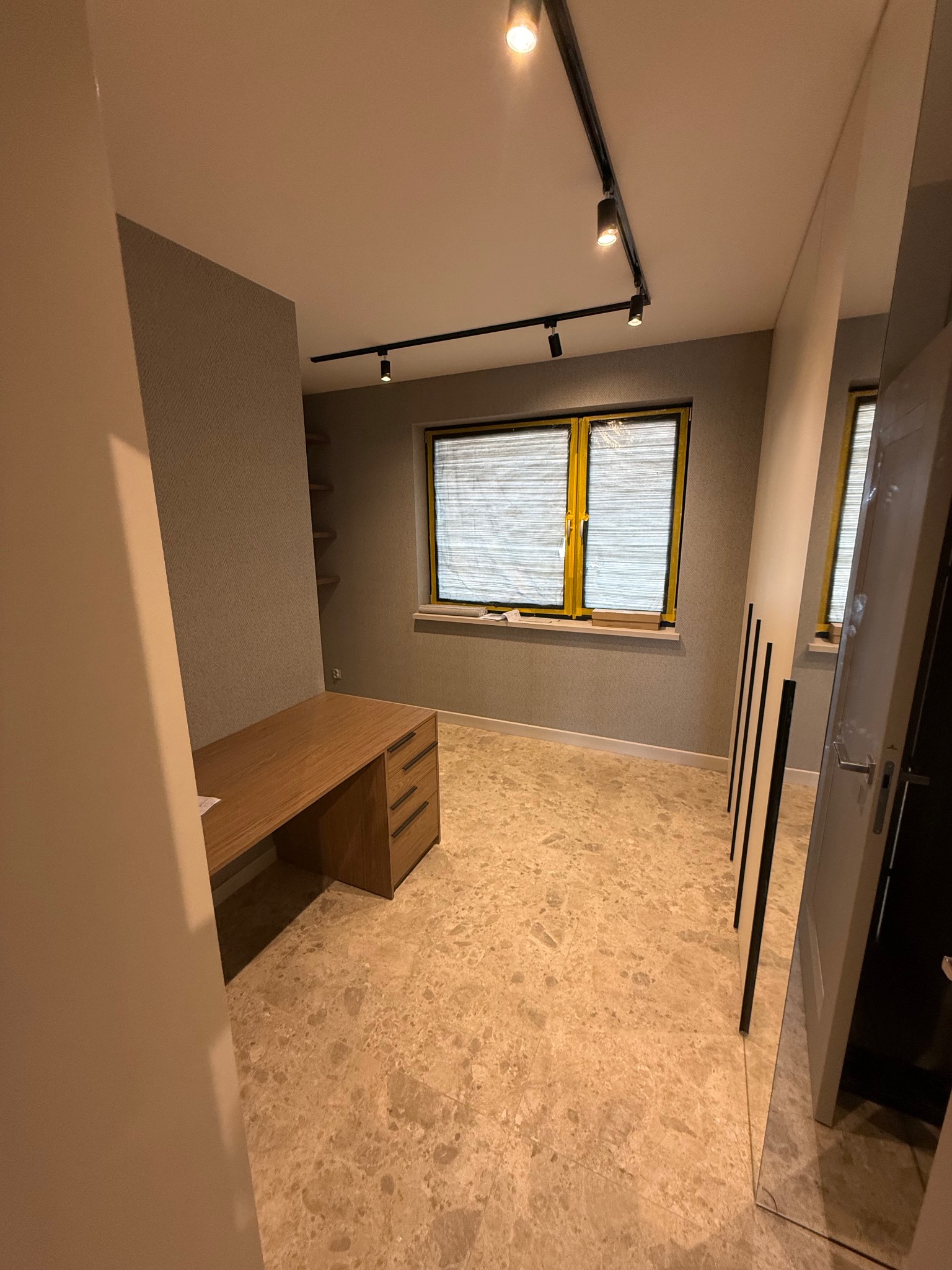 Jasne wnętrze pokoju po remoncie: marmurowa podłoga, biurko z drewna, okno z żółtymi ramami zabezpieczone folią, oświetlenie szynowe i lustro w drzwiach.