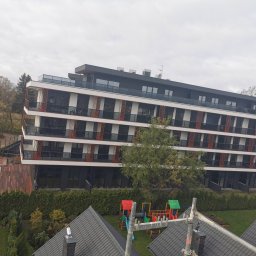 Jarbud - Nowoczesny apartamentowiec z balkonami i basenem, widok z góry. Na pierwszym planie fragment dachu i rusztowania, w tle drzewa i plac zabaw.
