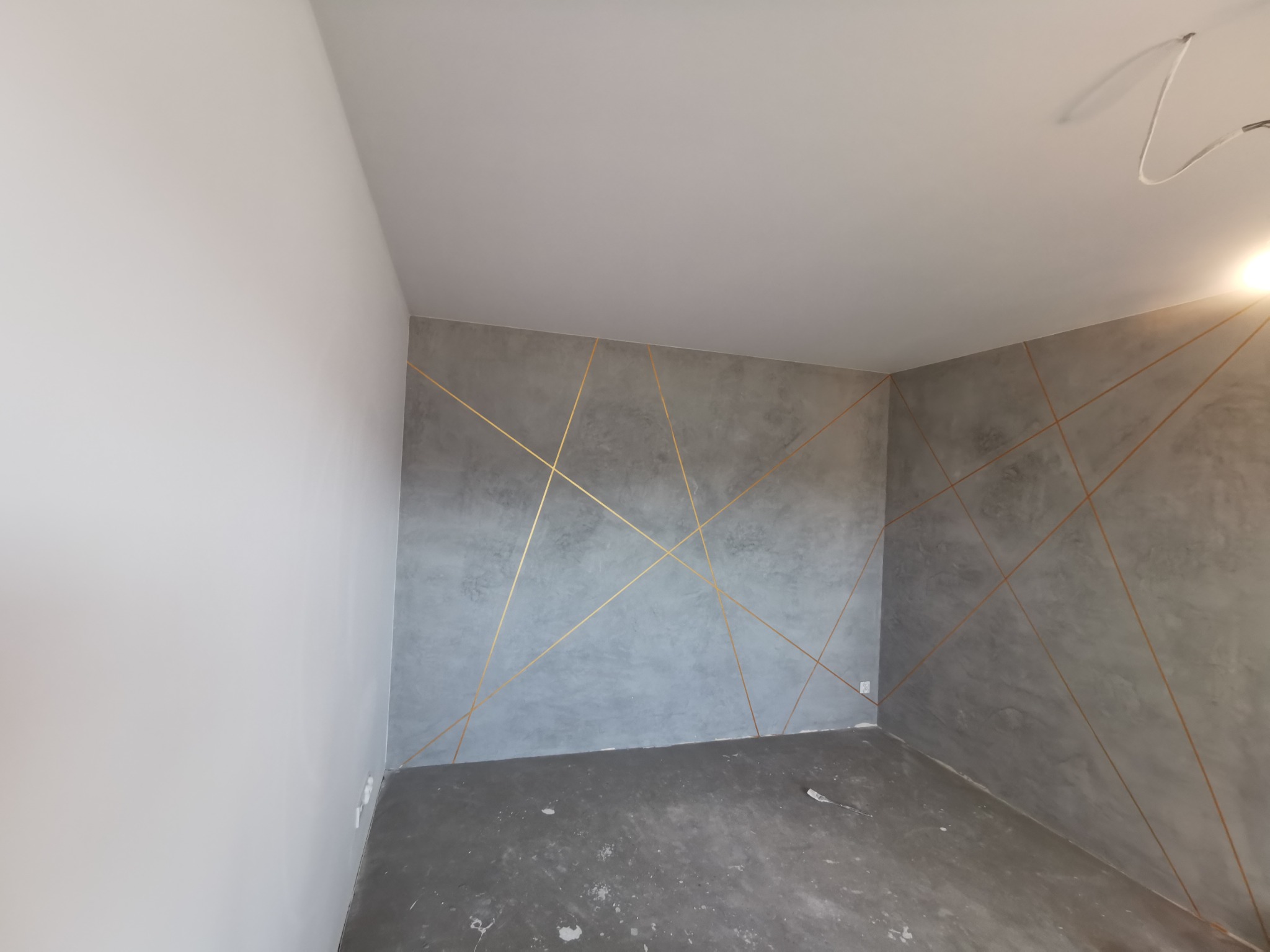 Szara ściana z dekoracyjnymi, złotymi liniami w pustym pokoju. Widoczne surowe wykończenie podłogi i fragment instalacji elektrycznej przy suficie.