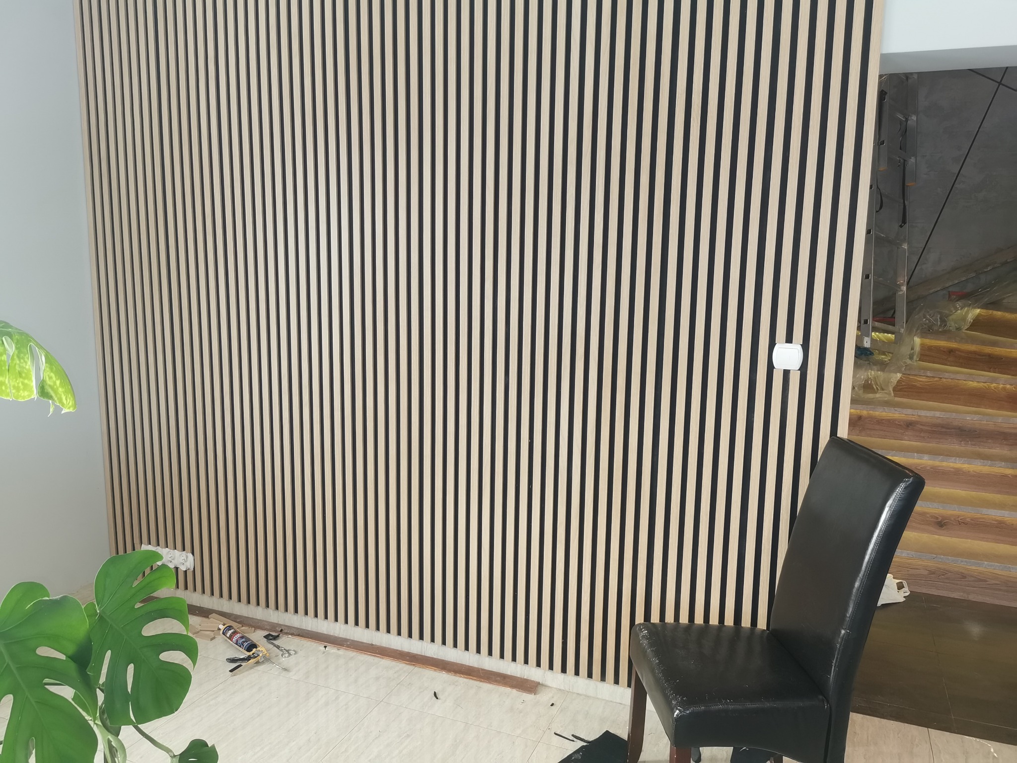 Ściana wykończona panelami lamelowymi w odcieniach beżu i czerni, z widocznym fragmentem krzesła i rośliny doniczkowej w dolnym rogu. Widoczne narzędzia i akcesoria montażowe.