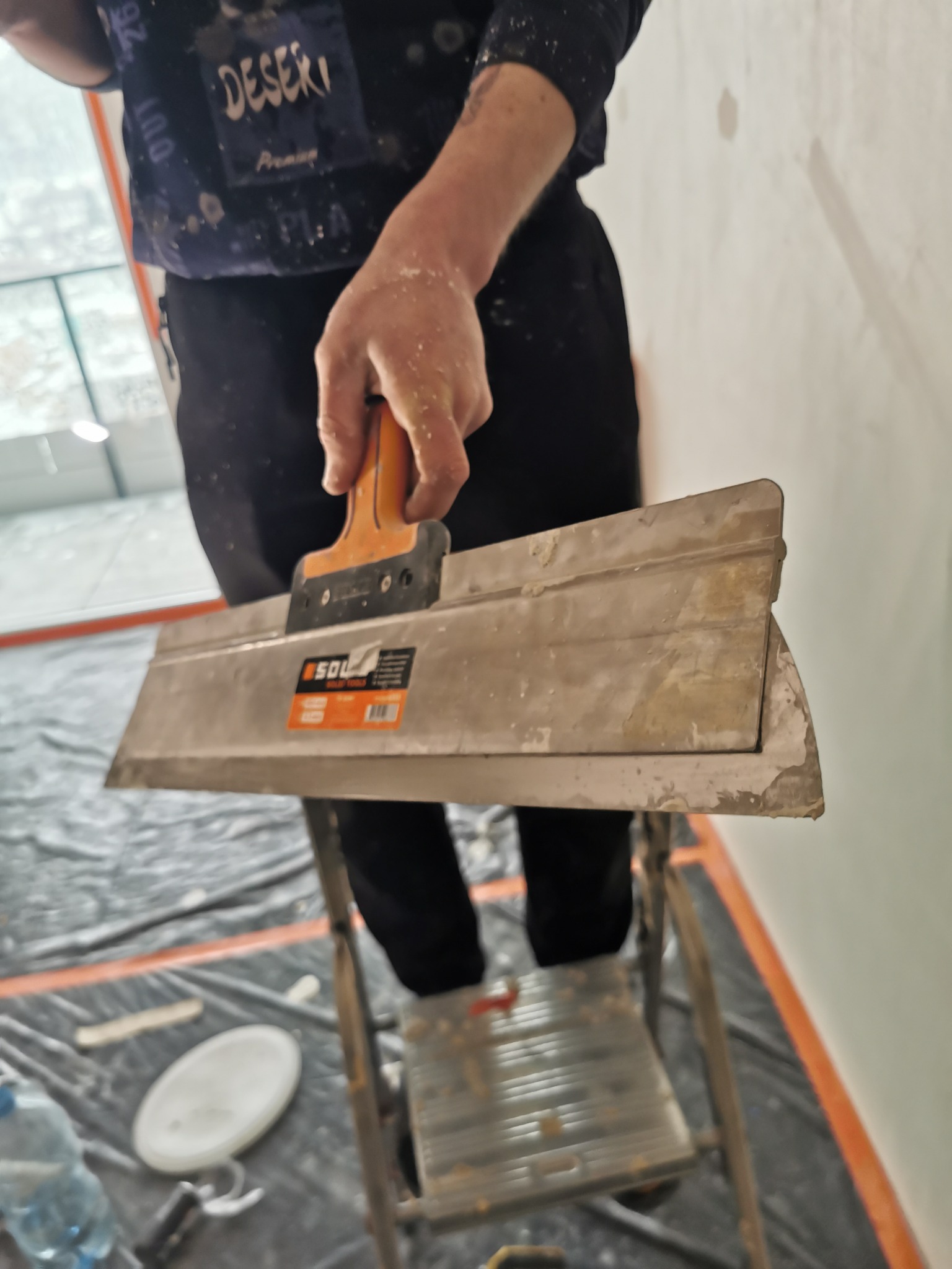 Pracownik zacierający gładź na ścianie za pomocą szerokiej szpachelki SOL, stojąc na drabinie. Podłoga zabezpieczona folią malarską.