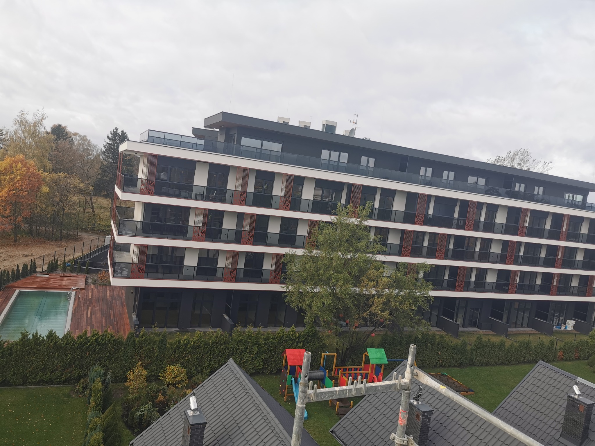 Nowoczesny apartamentowiec z balkonami i basenem, widok z góry. Na pierwszym planie fragment dachu i rusztowania, w tle drzewa i plac zabaw.